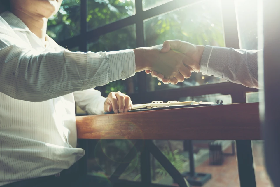 Deux professionnels en costume se serrent la main au-dessus d'une table en bois dans un bureau moderne avec baies vitrées